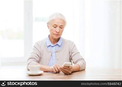 technology, communication age and people concept - senior woman with smartphone and coffee sitting at table and texting message at home