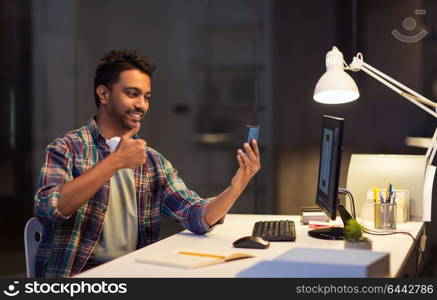 technology, communication and deadline concept - creative man with smartphone and computer having video call at night office and showing thumbs up. man with smartphone having video call at office