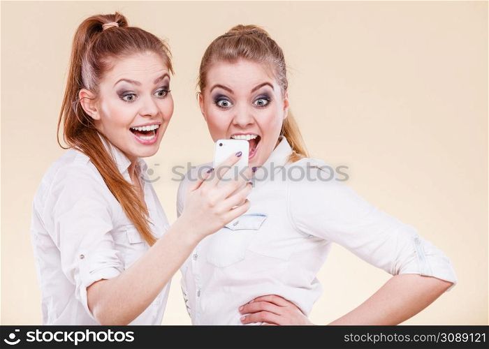 Technology communication and friendship concept. Two teen girls casual style using mobile phone reading message, funny surprised emotion on face. Girls using mobile phone reading message