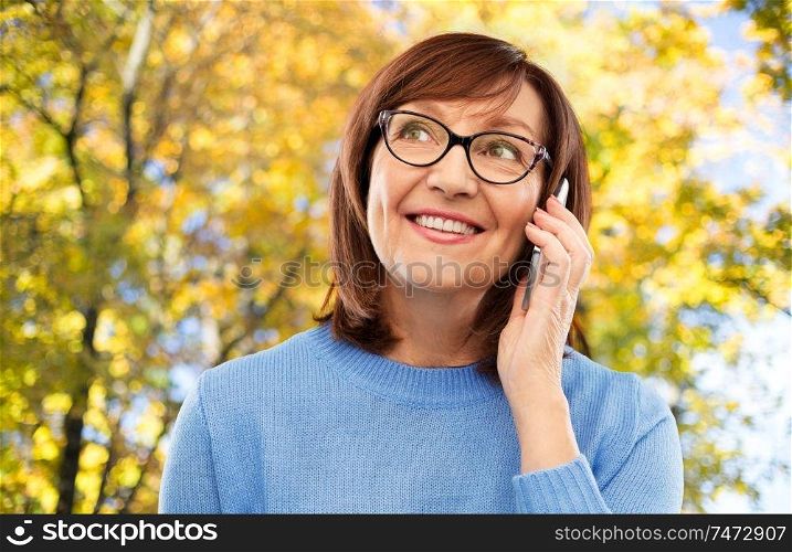 technology, communication and old people concept - portrait of smiling senior woman in glasses calling on smartphone over autumn park background. senior woman in glasses calling on smartphone