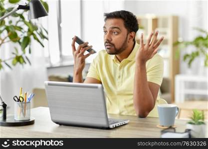 technology, communication and people concept - angry indian with smartphone and laptop computer using voice command recorder at home office. man recording voice on smartphone at home office