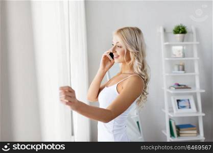 technology, communication and people concept - beautiful young woman in white underwear calling on smartphone and looking through window in morning. woman calling on smartphone at window in morning