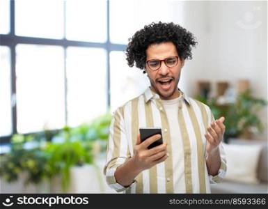 technology, communication and people concept - displeased man in glasses with smartphone over home room background. displeased young man with smartphone at home