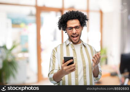 technology, communication and people concept - displeased man in glasses with smartphone over office background. displeased young man with smartphone at office