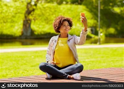 technology, communication and people concept - happy african woman taking selfie with smartphone and sitting on bench in summer park