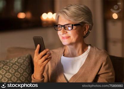 technology, communication and people concept - happy senior woman in glasses with smartphone at home in evening. happy senior woman with smartphone at home