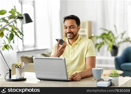 technology, communication and people concept - happy smiling indian with smartphone and laptop computer using voice command recorder at home office. man recording voice on smartphone at home office