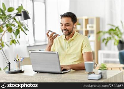 technology, communication and people concept - happy smiling indian with smartphone, earphones and laptop computer using voice command recorder at home office. man recording voice on smartphone at home office