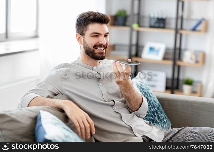 technology, communication and people concept - happy smiling man using voice command recorder on smartphone at home. happy man with smartphone recording voice at home