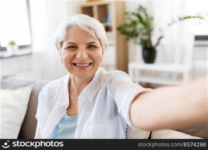 technology, communication and people concept - happy smiling senior woman in glasses taking selfie at home. smiling senior woman taking selfie at home