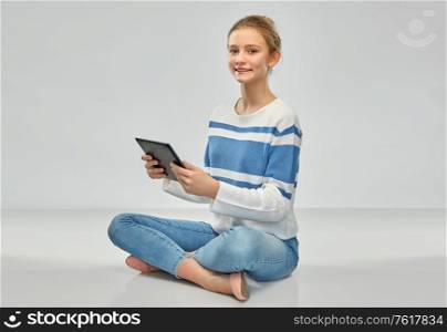 technology, communication and people concept - happy smiling teenage girl in pullover using tablet pc computer sitting on floor over grey background. happy smiling teenage girl using tablet computer