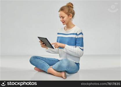 technology, communication and people concept - happy smiling teenage girl in pullover using tablet pc computer sitting on floor over grey background. happy smiling teenage girl using tablet computer
