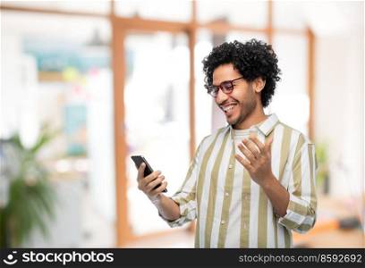 technology, communication and people concept - happy smiling young man in glasses with smartphone over office background. happy smiling young man with smartphone at office