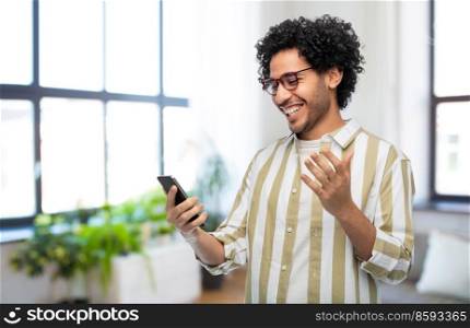 technology, communication and people concept - happy smiling young man in glasses with smartphone over home room background. happy smiling young man with smartphone at home