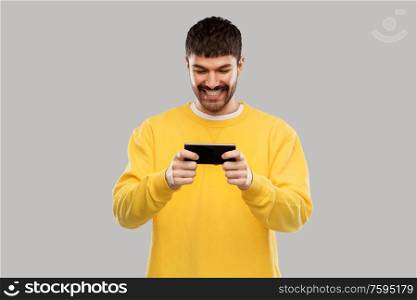 technology, communication and people concept - happy smiling young man playing game on smartphone over grey background. happy smiling young man with smartphone