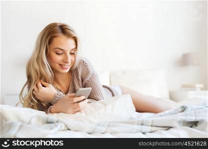 technology, communication and people concept - happy young woman lying in bed and texting on smartphone at home bedroom
