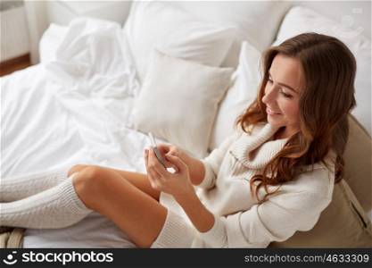 technology, communication and people concept - happy young woman lying in bed and texting on smartphone at home bedroom
