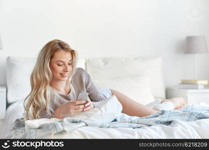 technology, communication and people concept - happy young woman lying in bed and texting on smartphone at home bedroom