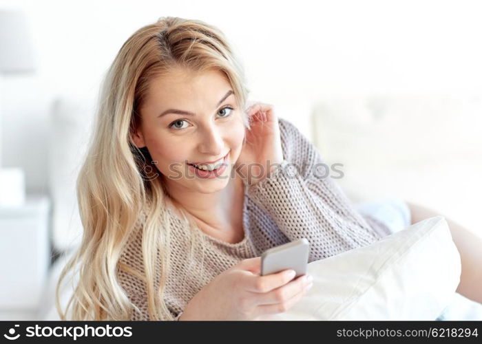 technology, communication and people concept - happy young woman lying in bed with smartphone at home bedroom