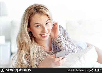 technology, communication and people concept - happy young woman lying in bed with smartphone at home bedroom