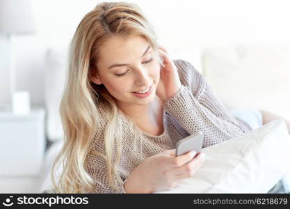 technology, communication and people concept - happy young woman lying in bed and texting on smartphone at home bedroom