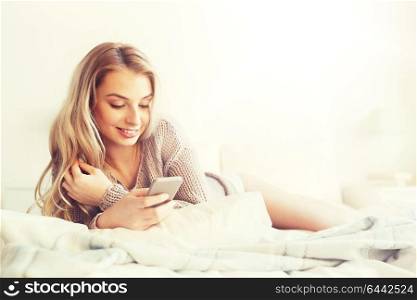 technology, communication and people concept - happy young woman lying in bed and texting on smartphone at home bedroom. happy young woman with smartphone in bed at home