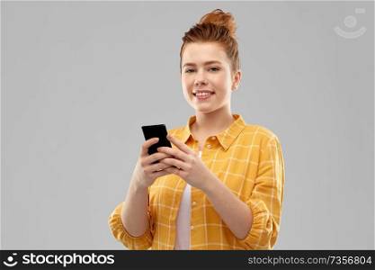 technology, communication and people concept - smiling red haired teenage girl in checkered shirt using smartphone over grey background. smiling red haired teenage girl using smartphone