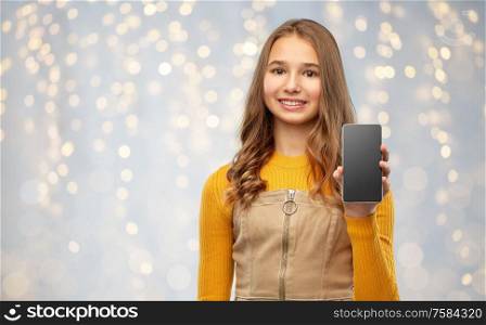 technology, communication and people concept - smiling teenage girl showing blank screen of smartphone over festive lights background. smiling teenage girl showing smartphone