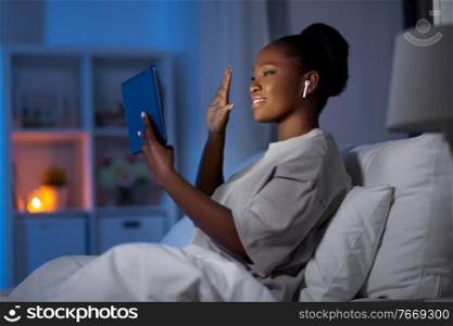 technology, communication and people concept - young african american woman with smartphone and earphones having video call in bed at home at night. woman with smartphone and earbuds in bed at night
