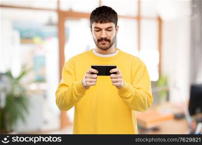 technology, communication and people concept - young man playing game on smartphone over office background. young man playing game on smartphone