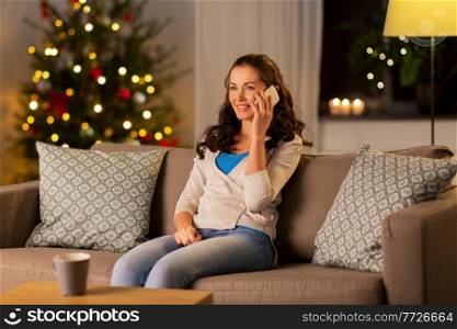 technology, communication and winter holidays concept - happy young woman calling on smartphone at home in evening over christmas tree lights on background. happy woman calling on phone at home on christmas