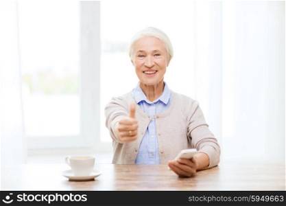 technology, communication, gesture, age and people concept - happy senior woman with smartphone and coffee sitting at table and showing thumbs up at home