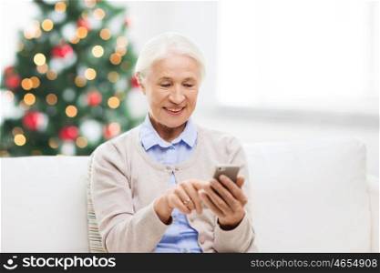 technology, communication, holidays age and people concept - happy senior woman with smartphone texting message at home over christmas tree lights background