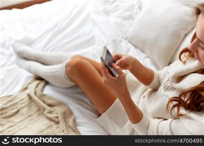 technology, communication, winter and people concept - happy young woman lying in bed and texting on smartphone at home bedroom
