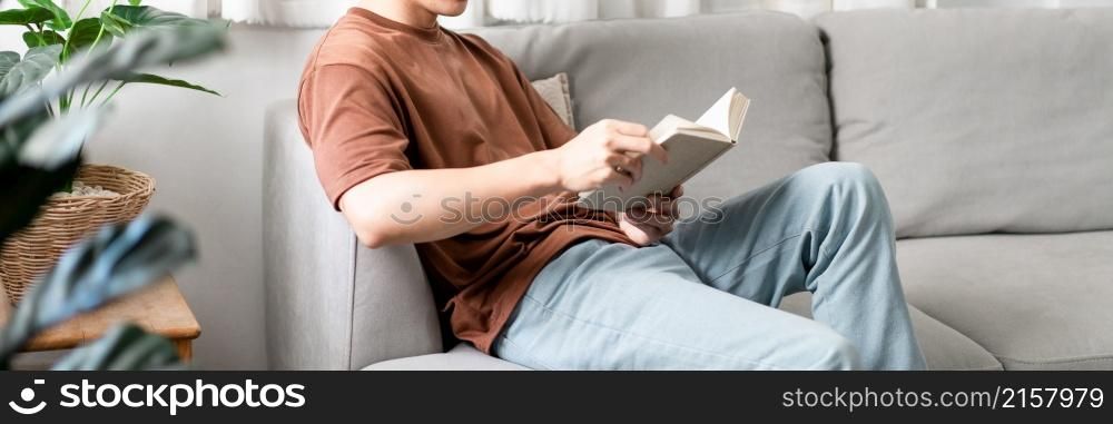 Technology Concept The male with his glasses sitting on the grey sofa, leaning his back on it, and reading a book.