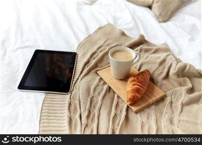 technology, coziness, morning and winter concept - tablet pc computer, coffee cup and croissant on bed at home