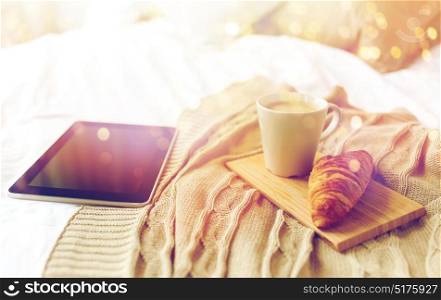 technology, coziness, morning and winter concept - tablet pc computer, coffee cup and croissant on bed at home. tablet pc, coffee and croissant on bed at home