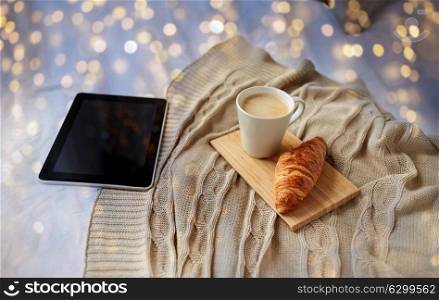 technology, coziness, morning and winter concept - tablet pc computer, coffee cup and croissant on bed at home. tablet pc, coffee and croissant on bed at home