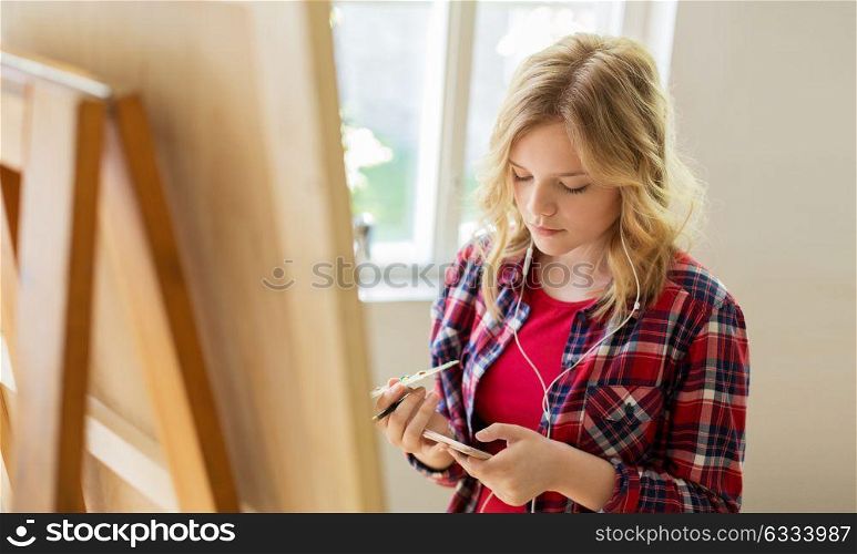 technology, creativity and people concept - teenage girl in earphones with smartphone and easel painting at art school studio. teenage girl with smartphone at art school studio