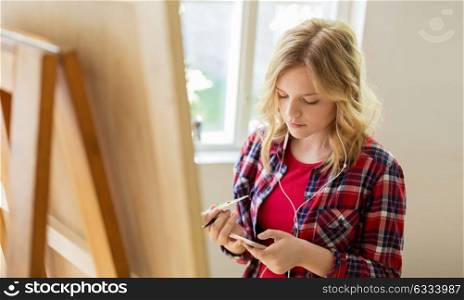 technology, creativity and people concept - teenage girl in earphones with smartphone and easel painting at art school studio. teenage girl with smartphone at art school studio