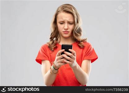 technology, cyberbullying and emotions concept - sad teenage girl in red t-shirt using smartphone over grey background. sad teenage girl using smartphone
