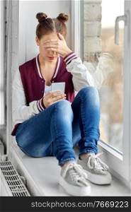 technology, cyberbullying and people concept - sad teenage girl with smartphone sitting on window sill. teenage girl with smartphone sitting on windowsill