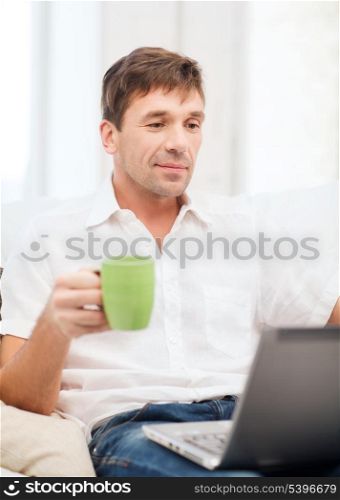 technology, drinks and lifestyle concept - man working with laptop at home, holding a cup of warm tea or coffee