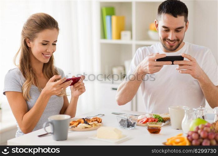 technology, eating and people concept - happy couple with smartphones having breakfast and photographing food at home. couple with smartphones having breakfast at home