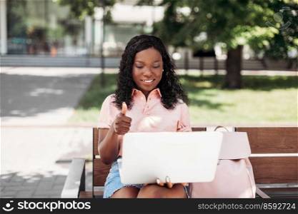 technology, education and peop≤concept - happy smiling african american student girl with laptop compute having video call and showing thumbs up in city. african student girl with laptop having video call