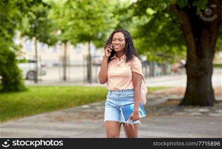 technology, education and people concept - happy smiling african american student girl with textbook calling on smartphone in city. african student girl calling on smartphone in city