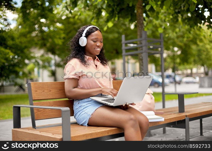 technology, education and people concept - happy smiling african american student girl in headphones with laptop computer and books in city. african student girl in headphones with laptop