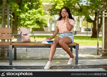 technology, education and people concept - happy smiling african american student girl with laptop computer and books calling on smartphone in city. african student girl calling on smartphone in city