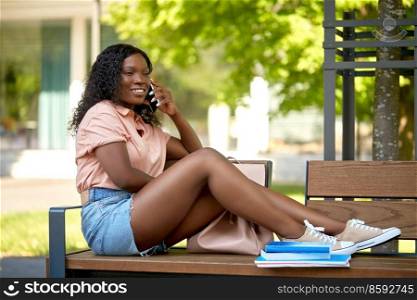 technology, education and people concept - happy smiling african american student girl with books calling on smartphone sitting on bench in city. african student girl calling on smartphone in city