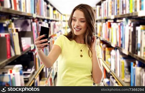 technology, education and people concept - smiling young woman or teenage girl in blank yellow t-shirt taking selfie by smartphone over book shelves in library background. teenage student girl taking selfie at library